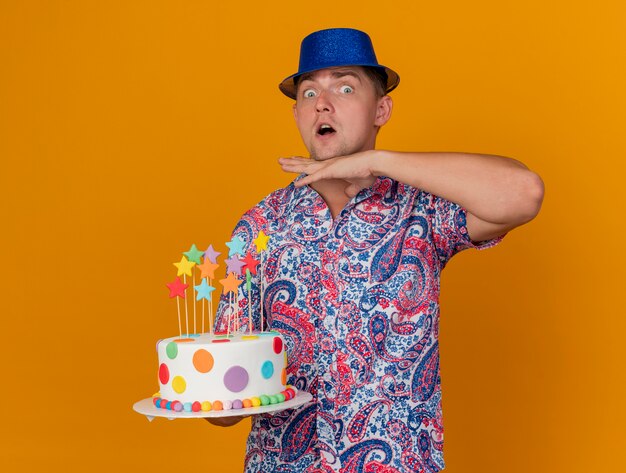 Jovem festeiro surpreso com um chapéu azul, segurando um bolo e colocando a mão na garganta isolada em laranja