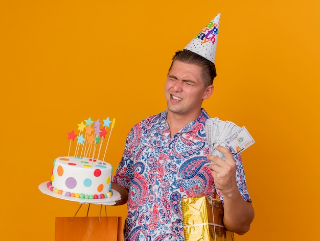 Jovem festeiro usando boné de aniversário com os olhos fechados segurando presentes com bolo e dinheiro isolado em laranja