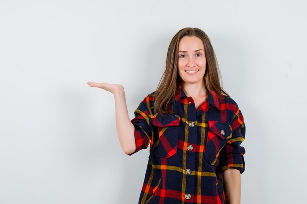 Jovem fingindo mostrar algo em uma camisa casual e olhando alegre, vista frontal.