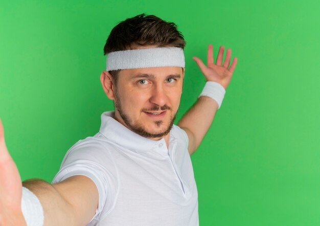 Jovem fitness com camisa branca e fita na cabeça, olhando para a frente, sorrindo amigável em pé sobre a parede verde