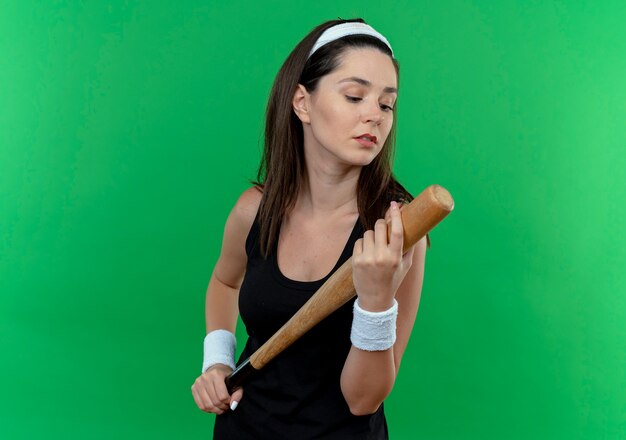 jovem fitness com uma bandana segurando um taco de beisebol olhando para ele com uma cara séria em pé sobre a parede verde