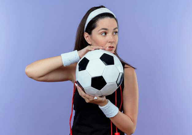 Jovem fitness com uma faixa na cabeça e pular corda no pescoço segurando uma bola de futebol, olhando para o lado com expressão confiante em pé sobre a parede azul