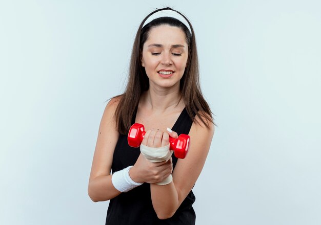 Jovem fitness com uma faixa na cabeça, malhando com halteres, parecendo confusa em pé sobre uma parede branca