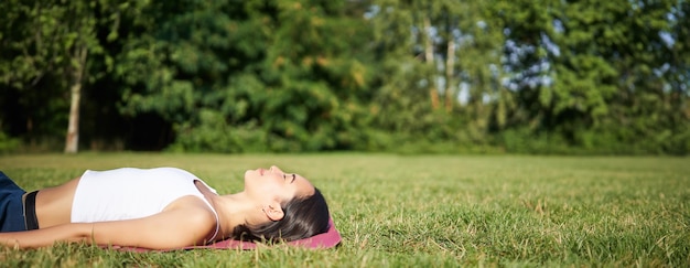 Foto grátis jovem fitness deitada no tapete esportivo no gramado, respirando e meditando no parque em roupas esportivas