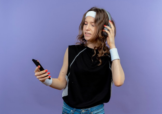 Jovem fitness em roupa esportiva preta com bandana e fones de ouvido, olhando para o smartphone, procurando música em pé sobre a parede azul