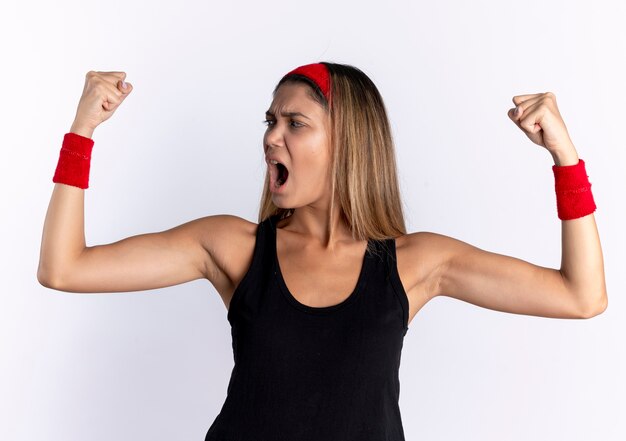 Jovem fitness garota em roupas esportivas pretas e bandana vermelha levantando os punhos e gritando com uma cara séria, gritando em pé sobre a parede branca
