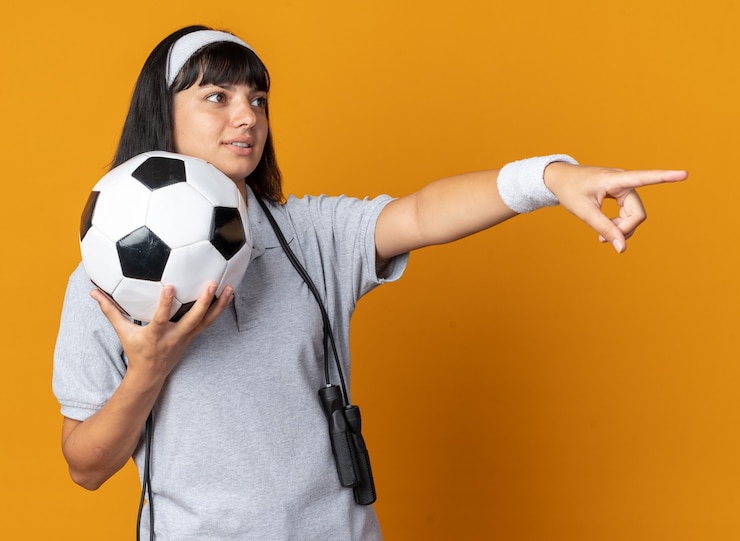 Foto grátis jovem fitness garota usando bandana e pular corda em volta do pescoço segurando uma bola de futebol, olhando para o lado com um sorriso no rosto apontando com o dedo indicador para algo em pé sobre um fundo laranja
