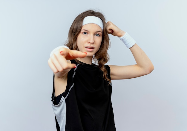 Jovem fitness girl em sportswear preto com tiara com expressão confiante apontando com o dedo levantando o punho mostrando bíceps em pé sobre uma parede branca