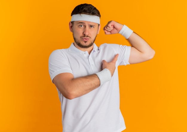 Jovem fitness homem com camisa branca e faixa na cabeça levantando o punho, mostrando os bíceps parecendo confiante e orgulhoso em pé sobre a parede laranja