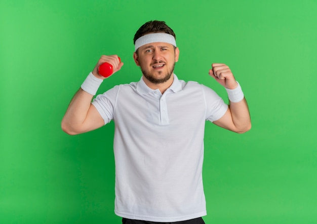 Foto grátis jovem fitness homem com camisa branca e faixa na cabeça, malhando com halteres tensos e confiante em pé sobre a parede verde