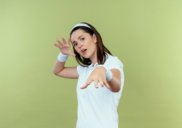 jovem fitness mulher com tiara fazendo gesto de defesa com as mãos e expressão de medo em pé sobre a parede de luz