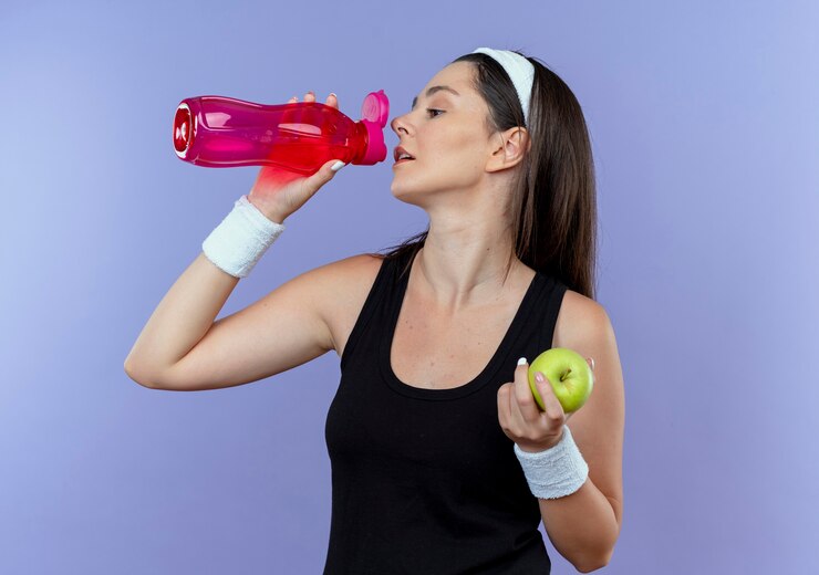 Foto grátis jovem fitness mulher com tiara segurando maçã verde bebendo água após o treino em pé sobre a parede azul