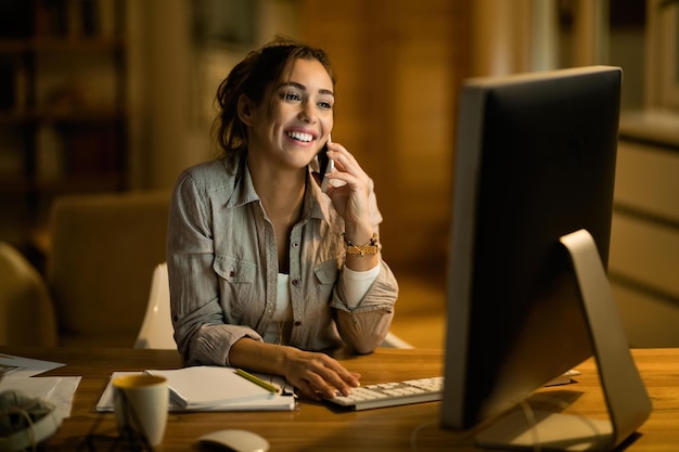 Jovem freelancer feliz se comunicando no celular enquanto navega na net no PC desktop à noite em casa