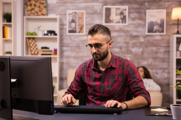 Foto grátis jovem freelancer focado enquanto trabalhava no computador de casa. namorada relaxando no sofá ao fundo.