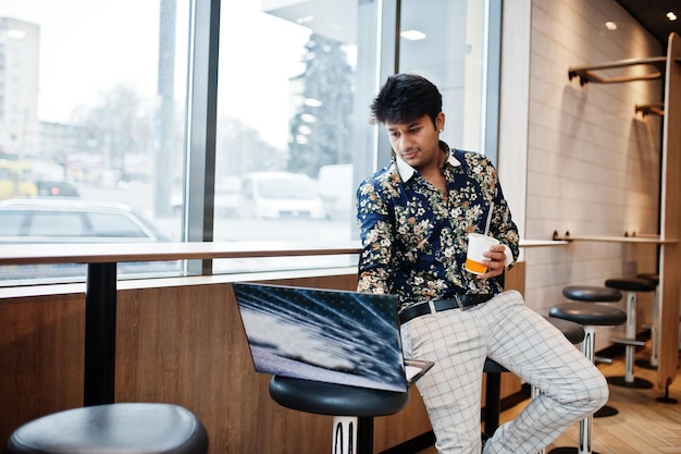 Jovem freelancer indiano masculino sentado computador portátil aberto no café de fast food homem asiático bonito lendo mensagem de texto durante o trabalho no netbook no café confortável