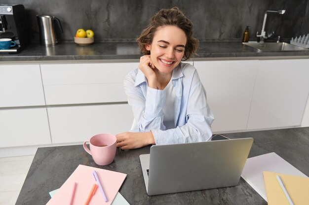 Jovem freelancer trabalha em casa, olha para a tela do laptop e sorri, faz lição de casa na cozinha