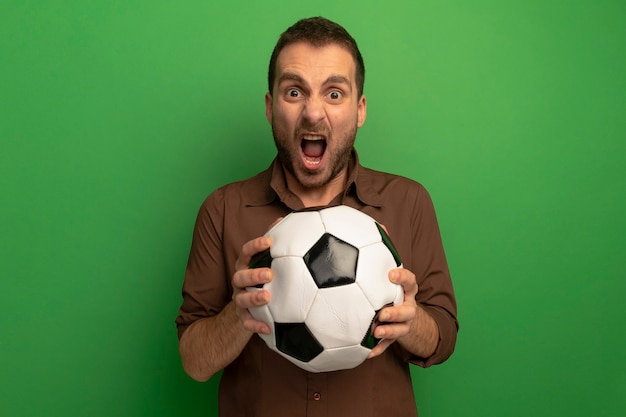 Jovem furioso segurando uma bola de futebol, olhando para a frente, gritando isolado na parede verde