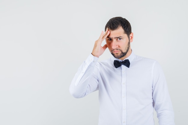 Jovem garçom de camisa branca com dor de cabeça e parecendo triste, vista frontal.