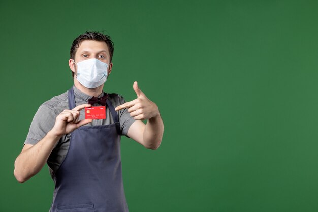 jovem garçom de uniforme com máscara médica e mostrando cartão do banco fazendo gesto de ok sobre fundo verde