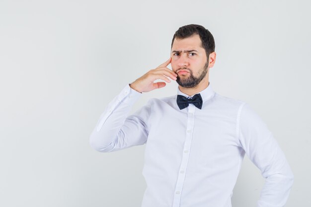 Jovem garçom em camisa branca em pé em pose de pensamento e olhando estrito, vista frontal.