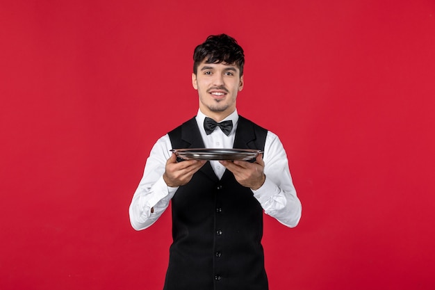 Jovem garçom satisfeito com um uniforme amarrando uma borboleta no pescoço segurando a bandeja no fundo vermelho isolado