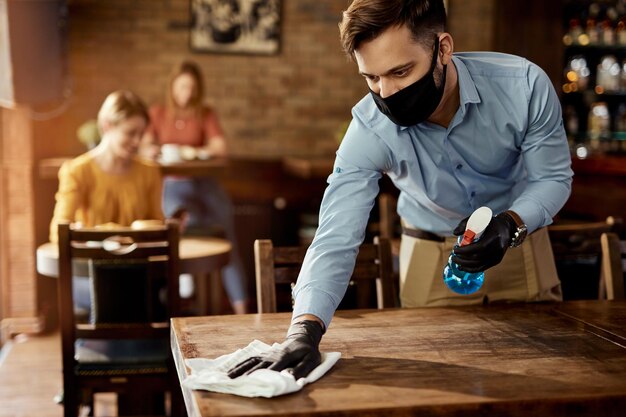 Jovem garçom usando máscara facial protetora enquanto limpa mesas enquanto trabalha em um café