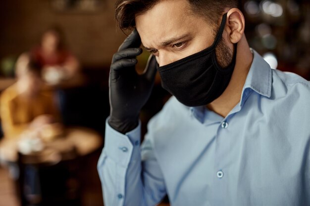Jovem garçom usando máscara facial protetora enquanto tem dor de cabeça no trabalho