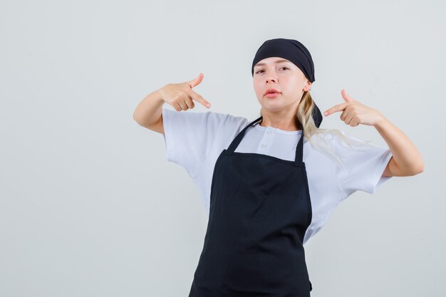 Jovem garçonete apontando para si mesma de uniforme e avental e parecendo orgulhosa