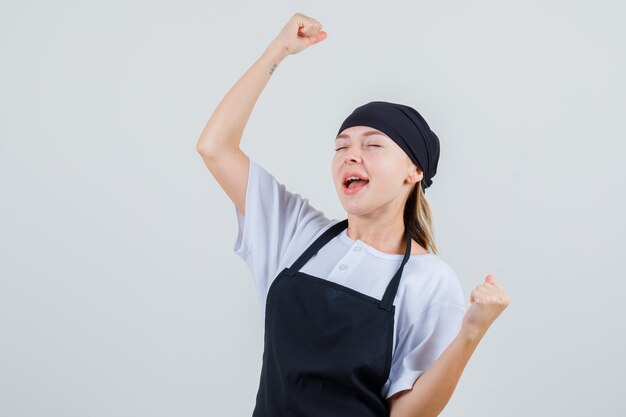 Jovem garçonete mostrando gesto de vencedor de uniforme e avental e parecendo feliz