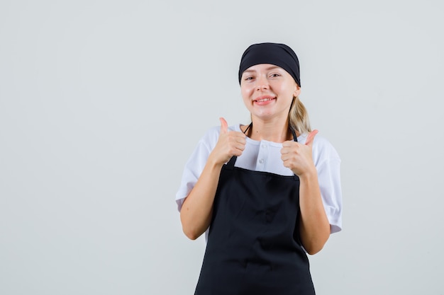 Foto grátis jovem garçonete mostrando o polegar para cima de uniforme e avental e parecendo satisfeita