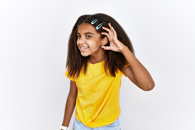 Foto grátis jovem garota afro-americana em pé sobre fundo branco isolado, sorrindo com a mão sobre a orelha, ouvindo uma audiência de rumores ou fofocas. conceito de surdez.