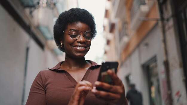 Jovem garota afro de óculos parecendo feliz mandando mensagens de texto com amigos