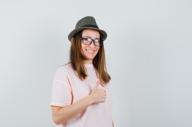 Jovem garota aparecendo o polegar em uma camiseta rosa, chapéu e olhando alegre, vista frontal.