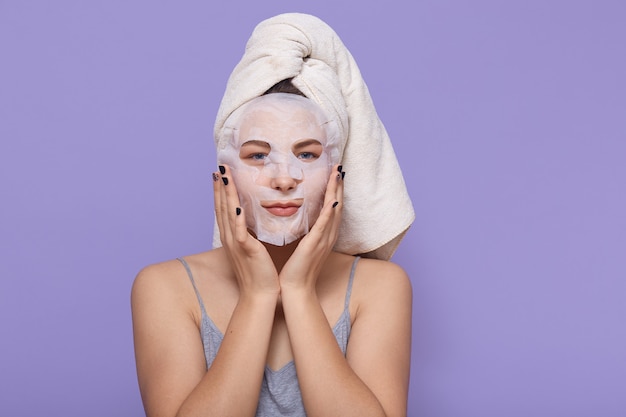 Jovem garota aplicar máscara facial, fazendo procedimentos de tratamento de beleza, vestindo uma toalha branca na cabeça