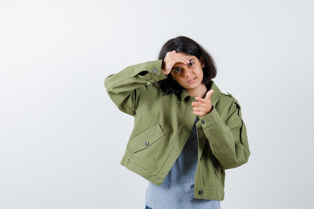 Jovem garota apontando enquanto segura a mão na testa em um suéter cinza, jaqueta cáqui, calça jeans e olhando sério. vista frontal.