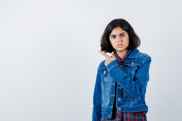 Jovem garota apontando para a esquerda com o dedo indicador em camisa e jaqueta jeans e olhando sério, vista frontal.