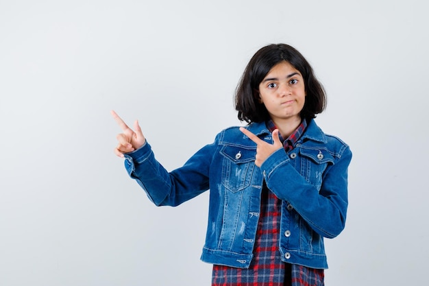 Jovem garota apontando para a esquerda com o dedo indicador em camisa xadrez e jaqueta jeans e parecendo irritada. vista frontal.