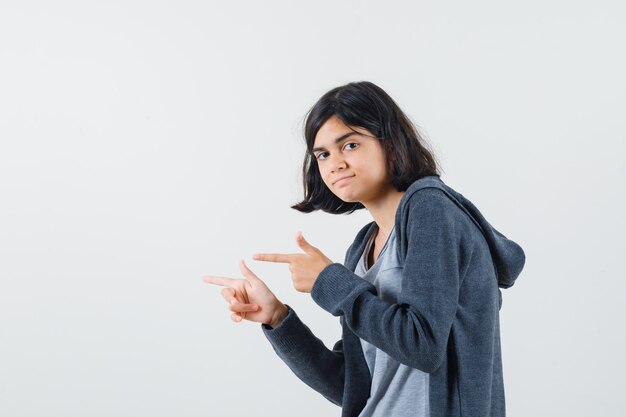 Jovem garota apontando para a esquerda com os dedos indicadores em uma camiseta cinza claro e um capuz cinza escuro com zíper na frente e parecendo séria.