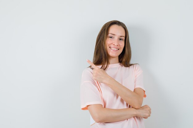 Jovem garota apontando para o canto superior esquerdo em uma camiseta rosa e olhando alegre, vista frontal.