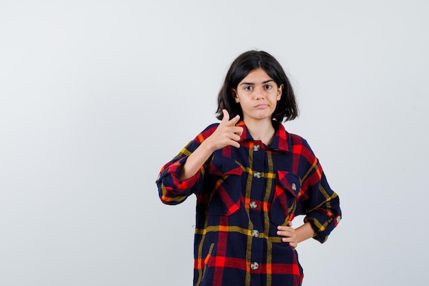 Jovem garota apontando, segurando a mão na cintura em uma camisa quadriculada e olhando bonita, vista frontal.