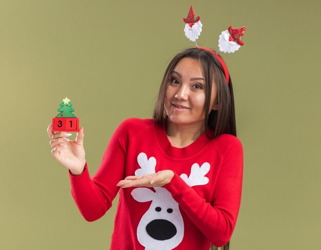 Foto grátis jovem garota asiática satisfeita vestindo uma argola de cabelo de natal segurando e apontando com a mão para um brinquedo de natal isolado em fundo verde oliva