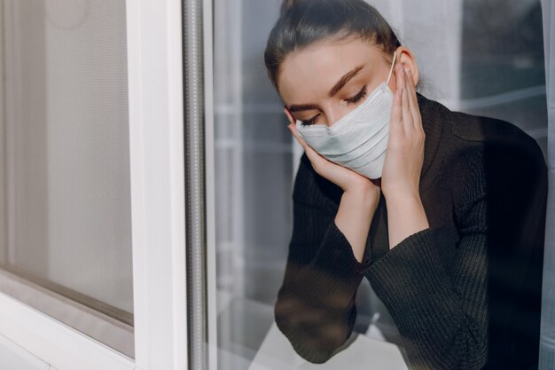Jovem garota atraente em uma máscara médica protetora olha pela janela. isolamento durante a epidemia. isolamento em casa.
