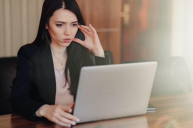 Jovem garota atraente emocional em roupas de estilo empresarial, sentado em uma mesa em um laptop e telefone no escritório ou auditório