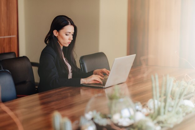 Jovem garota atraente emocional em roupas de estilo empresarial, sentado em uma mesa em um laptop e telefone no escritório ou auditório