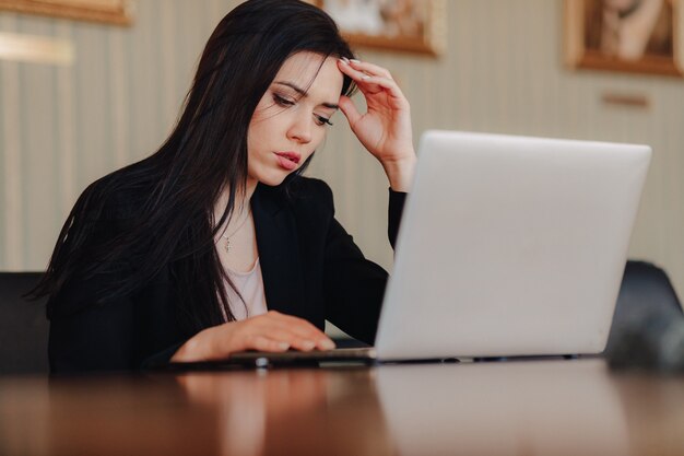 Jovem garota atraente emocional em roupas de estilo empresarial, sentado em uma mesa em um laptop e telefone no escritório ou auditório