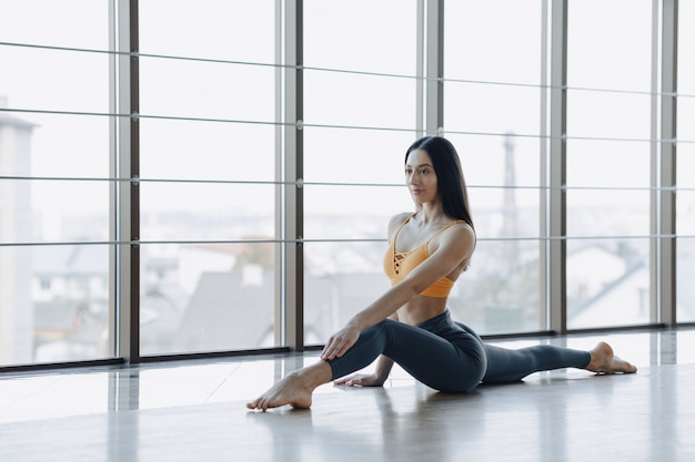 Jovem garota atraente fazendo exercícios de fitness com yoga no chão contra a superfície das janelas panorâmicas