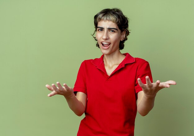 Jovem garota brava caucasiana com corte de cabelo de duende mostrando as mãos vazias isoladas em um fundo verde oliva com espaço de cópia