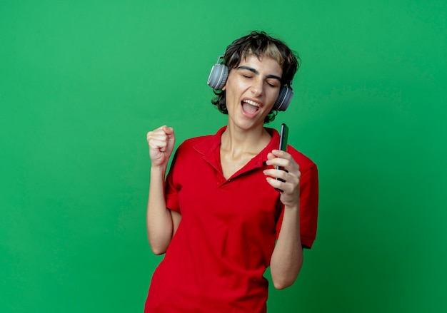 Jovem garota caucasiana com corte de cabelo de duende usando fones de ouvido, ouvindo música, segurando o telefone celular, fingindo cantar usando o telefone como microfone com o punho cerrado e olhos fechados