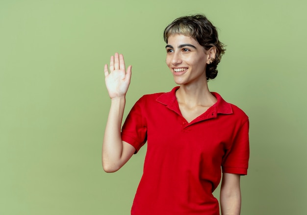 Jovem garota caucasiana sorridente com corte de cabelo de duende olhando para o lado fazendo gesto de olá isolado em fundo verde oliva com espaço de cópia