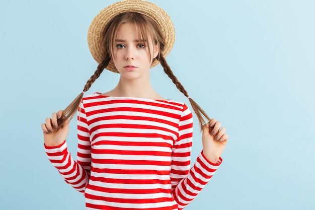 Foto grátis jovem garota chateada com duas tranças no chapéu de palha e colete listrado vermelho olhando tristemente na câmera enquanto passa o tempo sobre fundo azul
